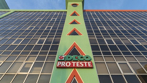 Plano de baixo para cima com vista parcial da fachada do edifício sede da DECO PROTESTE. A fachada é composta por várias fileiras contíguas de janelas, e ao centro um pilar de betão, pintado com as cores verde e vermelha, sobre o qual assenta o logótipo da marca, também ele decorado com as cores verde e vermelha.